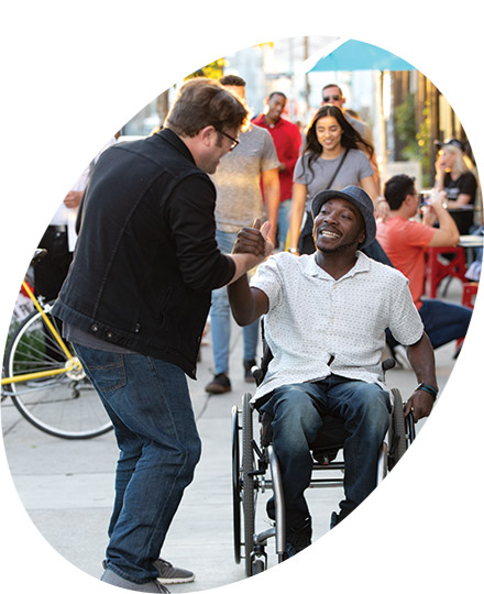 Man in wheelchair smiling and clasping hands with another man on the street